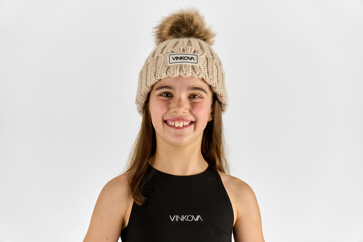 Beige Vinkova Wintermütze aus Strick mit Bommel an einer Turnerin.